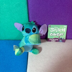 May include: A blue and teal plush Stitch character with large purple ears and black eyes. Beside it is a square green collection sheet with the text Disney Stitch Cutie Cuff Collection Sheet and a small illustration of the character.
