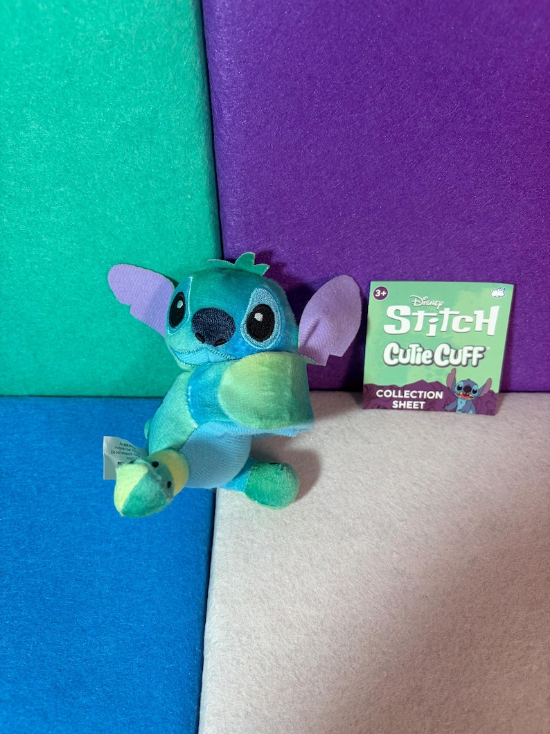 May include: A blue and teal plush Stitch character with large purple ears and black eyes. Beside it is a square green collection sheet with the text Disney Stitch Cutie Cuff Collection Sheet and a small illustration of the character.