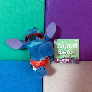 Puede incluir: Un peluche azul del personaje Stitch con gafas de sol rojas y pantalones cortos rojos con patrones florales blancos. Junto al peluche hay una tarjeta cuadrada de papel verde con el texto Disney Stitch Cutie Cuff COLLECTION SHEET.
