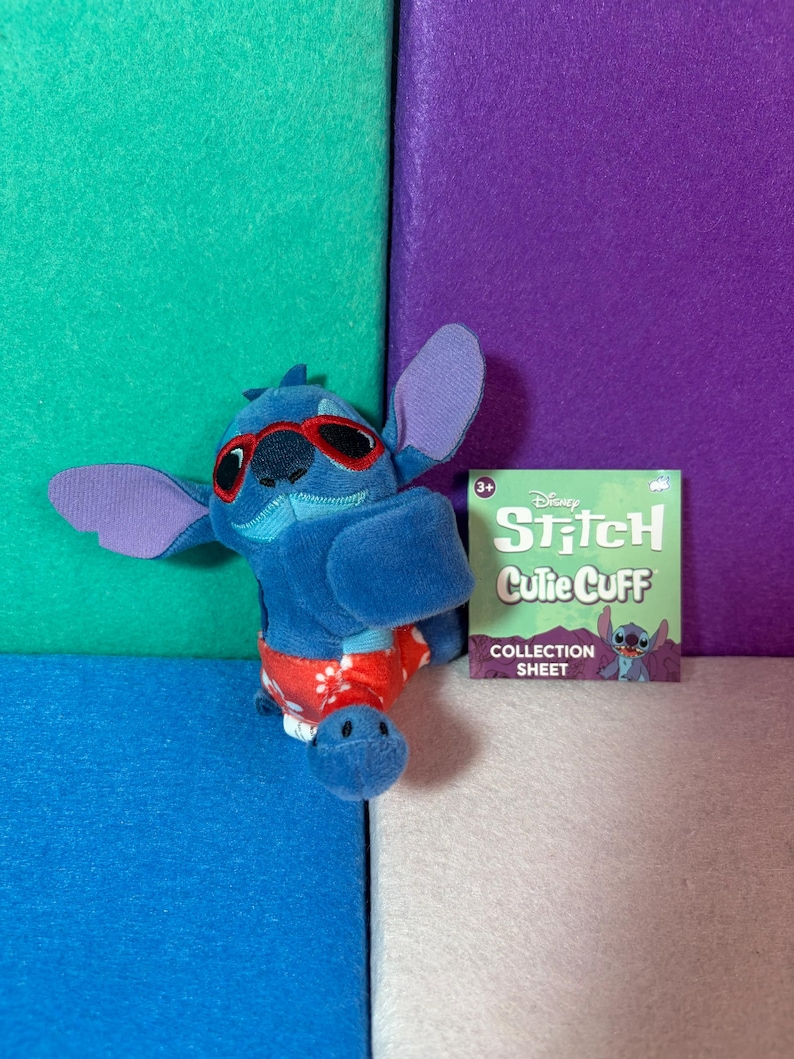 Puede incluir: Un peluche azul del personaje Stitch con gafas de sol rojas y pantalones cortos rojos con patrones florales blancos. Junto al peluche hay una tarjeta cuadrada de papel verde con el texto Disney Stitch Cutie Cuff COLLECTION SHEET.