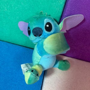 May include: A small plush toy character featuring a blue and light green color scheme with large ears, a dark blue nose, and embroidered eyes. The character has a soft texture and a white fabric tag attached to the side, displaying visible text.