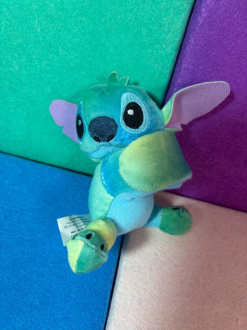 May include: A small plush toy character featuring a blue and light green color scheme with large ears, a dark blue nose, and embroidered eyes. The character has a soft texture and a white fabric tag attached to the side, displaying visible text.