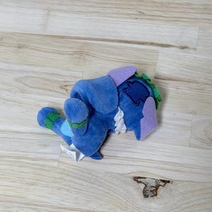 May include: A blue plush toy featuring a soft texture, light purple ears, and green accents on the feet and head. The character has a white ruffled collar detail and a dark blue patch on the back, with a small white fabric tag attached to the side.