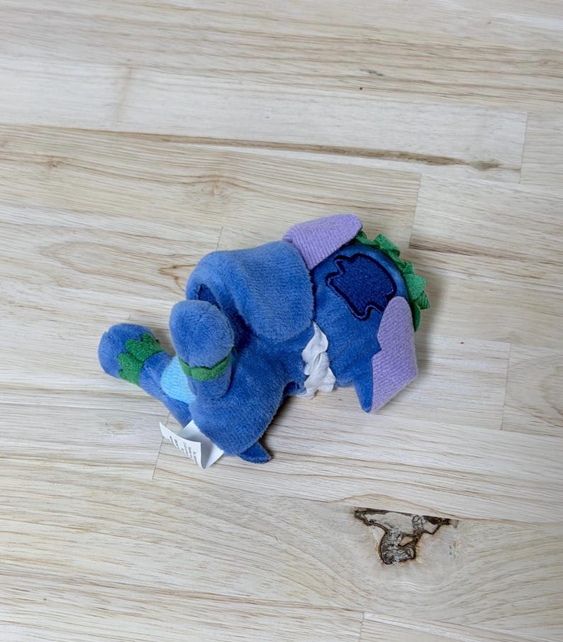 May include: A blue plush toy featuring a soft texture, light purple ears, and green accents on the feet and head. The character has a white ruffled collar detail and a dark blue patch on the back, with a small white fabric tag attached to the side.