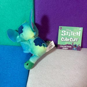 May include: A plush Stitch character toy featuring a blue and green gradient color scheme with dark blue patches on the back and ears. Beside it is a square green collection sheet with the text Disney Stitch Cutie Cuff Collection Sheet.