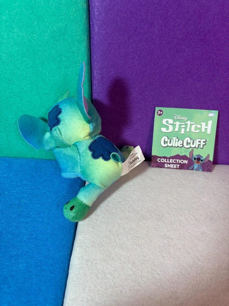 May include: A plush Stitch character toy featuring a blue and green gradient color scheme with dark blue patches on the back and ears. Beside it is a square green collection sheet with the text Disney Stitch Cutie Cuff Collection Sheet.
