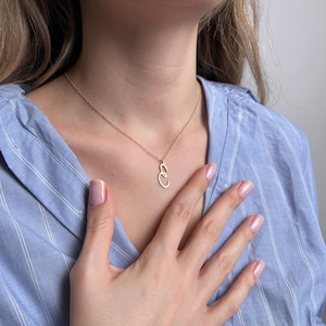 May include: A delicate gold necklace with a pendant featuring two intertwined oval shapes. The necklace is worn against a blue striped shirt. The pendant is approximately 0.75 inches in length.