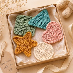 Puede incluir: Una caja de artículos de ganchillo hechos a mano en varias formas y colores. Incluye un cuadrado verde, un triángulo turquesa, un corazón rosa, una estrella de mar amarilla y un círculo crema. La caja también contiene una etiqueta que dice "handmade with love".