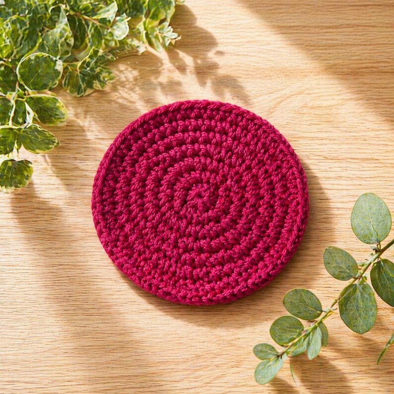 May include: A round, crocheted coaster in a deep magenta color. The coaster is made of thick yarn and features a spiral pattern. The coaster is placed on a light-colored wooden surface, with green foliage in the background.
