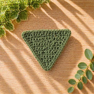 May include: A handmade, green crochet coaster in a triangular shape. The coaster is made with a textured stitch and has a defined border. The coaster is displayed on a wooden surface with green foliage in the background.