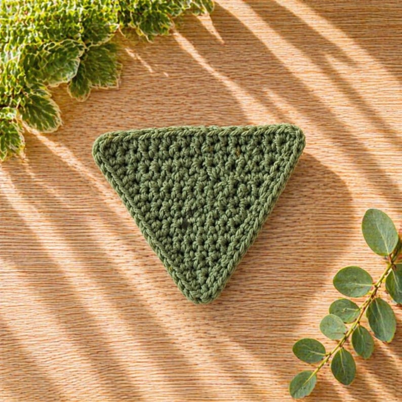 May include: A handmade, green crochet coaster in a triangular shape. The coaster is made with a textured stitch and has a defined border. The coaster is displayed on a wooden surface with green foliage in the background.