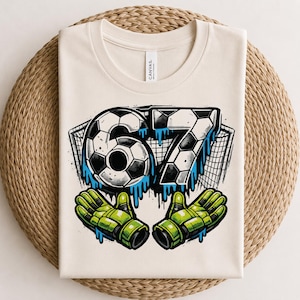 Puede incluir: Una camiseta color crema con un gráfico del número 67 diseñado con un patrón de balón de fútbol y efectos de pintura azul que gotea. Detrás del número hay una red de portería y debajo dos guantes de portero de color verde brillante.