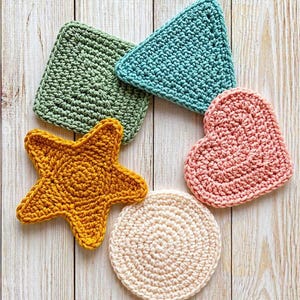 May include: A collection of handmade crochet coasters in various shapes and colors. Includes a square green coaster, a triangular teal coaster, a starfish-shaped yellow coaster, a heart-shaped pink coaster, and a round cream-colored coaster. The coasters are arranged on a light wood surface.
