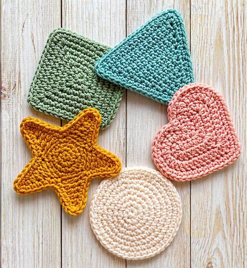 May include: A collection of handmade crochet coasters in various shapes and colors. Includes a square green coaster, a triangular teal coaster, a starfish-shaped yellow coaster, a heart-shaped pink coaster, and a round cream-colored coaster. The coasters are arranged on a light wood surface.