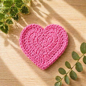 May include: A handmade, pink crochet heart coaster. The coaster is made of thick yarn and has a textured appearance. The heart shape is centered on a light wooden surface, with green leafy sprigs in the corners.