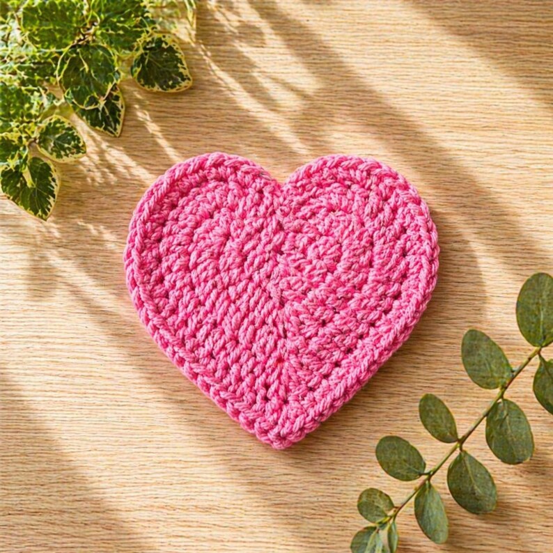 May include: A handmade, pink crochet heart coaster. The coaster is made of thick yarn and has a textured appearance. The heart shape is centered on a light wooden surface, with green leafy sprigs in the corners.