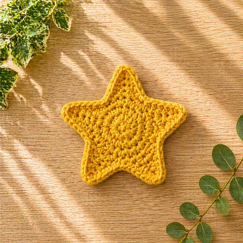May include: A handmade, yellow crochet star coaster. The star is made with a tight stitch pattern and has a textured appearance. The coaster is placed on a wooden surface, with green foliage in the background.