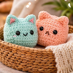 Puede incluir: Dos juguetes de gato hechos a mano con ganchillo en una cesta de mimbre. Un gato es de color azul claro con detalles rosas en las orejas y una cara sonriente. El otro es de color melocotón con una cara fruncida. Ambos tienen ojos de botón negros.