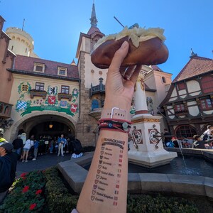 May include: A baked potato topped with a white substance is held up in front of a building with a tall spire. The arm holding the potato has a tattoo that says "Snacking" with a list of countries and flags.