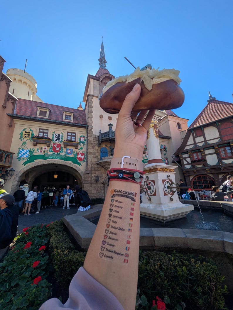 May include: A baked potato topped with a white substance is held up in front of a building with a tall spire. The arm holding the potato has a tattoo that says "Snacking" with a list of countries and flags.