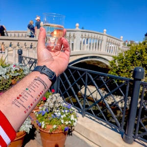 Op de afbeelding: Een doorzichtige plastic beker in een hand met een tijdelijke tatoeage op de onderarm. De tatoeage bevat de tekst Drinking Around The World, een lijst met landen waaronder Mexico, Noorwegen, China, Duitsland, Itali&euml;, Verenigde Staten, Marokko, Frankrijk, Verenigd Koninkrijk en Canada, elk met een klein Mickey Mouse-icoontje en een bijbehorende nationale vlag.