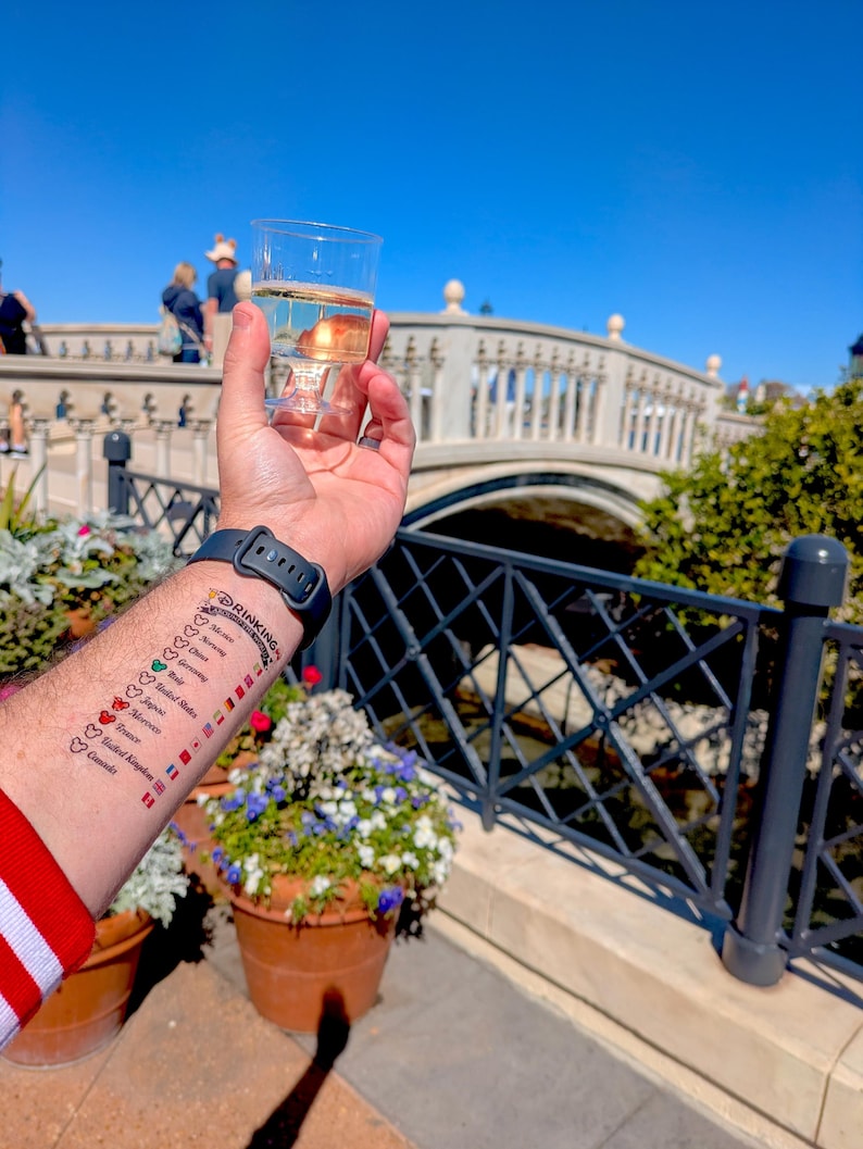 Op de afbeelding: Een doorzichtige plastic beker in een hand met een tijdelijke tatoeage op de onderarm. De tatoeage bevat de tekst Drinking Around The World, een lijst met landen waaronder Mexico, Noorwegen, China, Duitsland, Itali&euml;, Verenigde Staten, Marokko, Frankrijk, Verenigd Koninkrijk en Canada, elk met een klein Mickey Mouse-icoontje en een bijbehorende nationale vlag.
