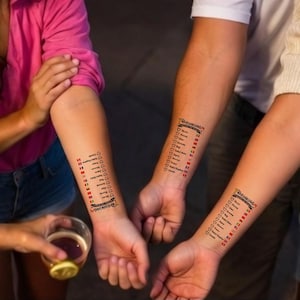 Op de afbeelding: Drie tijdelijke tatoeages op onderarmen, elk met een verticale lijst getiteld "DRINKING AROUND THE WORLD" met landnamen, bijbehorende nationale vlaggen en lege hartpictogrammen voor het bijhouden van de consumptie tijdens evenementen.