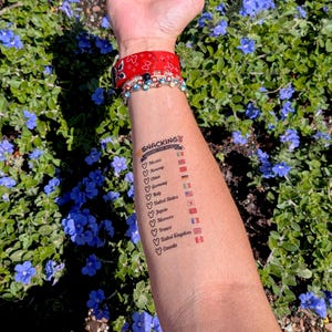 May include: A temporary tattoo on an arm lists countries with flags and the text "Snacking Around the World." The arm also has a red patterned wristband and multiple bracelets. The background features green foliage and blue flowers.