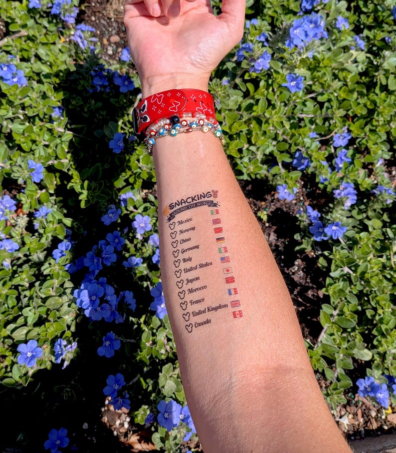 May include: A temporary tattoo on an arm lists countries with flags and the text "Snacking Around the World." The arm also has a red patterned wristband and multiple bracelets. The background features green foliage and blue flowers.