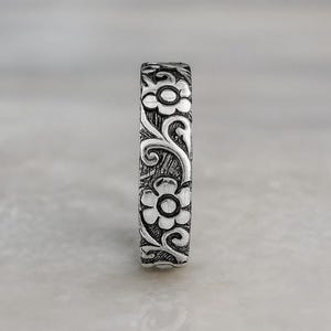 Op de afbeelding: Een zilveren ring met een bloemmotief. De ring heeft een herhalend ontwerp van bloemen en wervelende ranken. De ring is gemaakt van een glanzend, metalen materiaal en wordt waarschijnlijk om een vinger gedragen. De ring is een sieraad.