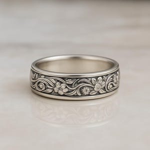May include: A silver ring with a floral pattern. The ring features a detailed design of flowers and vines around the band. The ring is displayed on a reflective surface.