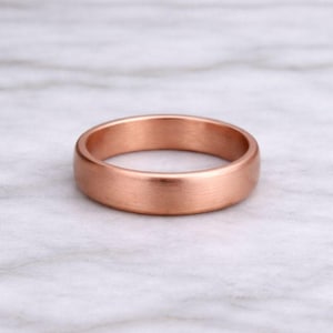 Op de afbeelding: Een roségouden ring met een geborstelde afwerking. De ring heeft een gladde, afgeronde band. De ring wordt gepresenteerd op een wit en grijs marmeren oppervlak. Deze ring is een sieraad.