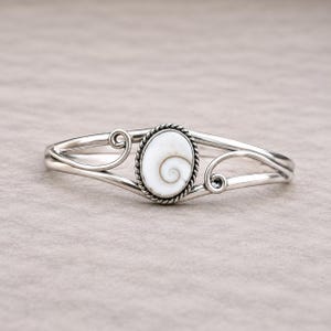 May include: A silver cuff bracelet featuring a central oval-shaped white shiva eye shell with a natural spiral pattern. The shell is set within a twisted silver border, flanked by polished silver bands that curve into decorative scroll accents.