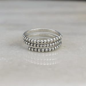 May include: A silver-toned spiral ring with a textured beaded design. The ring features three bands of small, evenly spaced beads that wrap around the finger. The ring is displayed on a light-colored surface.