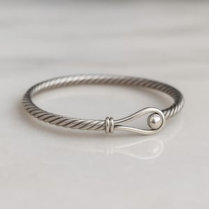 May include: A silver bangle bracelet with a twisted rope design. The bracelet has a hook-and-eye closure with a small, round ball. The metal has a polished finish, reflecting light.