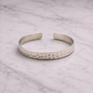 May include: A silver cuff bracelet with a hammered texture. The open-ended bracelet is displayed on a light grey marble surface. The bracelet's width is approximately 1.3 cm.