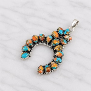 Può includere: Un pendente in argento a forma di ferro di cavallo, ornato da numerose pietre a forma di goccia. Le pietre presentano un mix di tonalità turchese, arancione e marrone, su sfondo bianco. Il pendente ha un'ansa in argento.