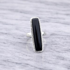 Könnte beinhalten: Ein Silberring mit einem langen, rechteckigen schwarzen Onyxstein. Der Stein ist in einer silbernen Lünette mit strukturiertem Rand gefasst. Der Ring wird auf einer hellgrauen Marmoroberfläche präsentiert.