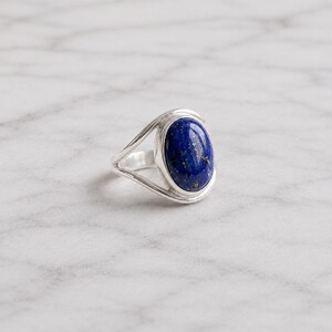 Può includere: Un anello in argento con una pietra preziosa di lapislazzuli lucida di forma ovale con visibili pagliuzze di pirite color oro. La fascia è composta da due fili d'argento lisci e sottili che convergono alla base dell'incastonatura.