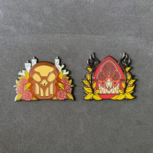 Könnte beinhalten: Zwei Emaille-Anstecknadeln. Eine Anstecknadel zeigt ein Schädeldesign mit Kerzen und Rosen in Orange-, Gelb- und Rottönen. Die andere Anstecknadel hat ein gotisches Bogendesign mit floralen und Geweihmotiven in Rot, Gelb und Braun.