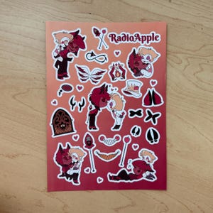 May include: A sticker sheet with various cartoon illustrations in red, black, and white. The sheet features characters and symbols, with the text "RadioApple" at the top. The background has a gradient from red to orange.