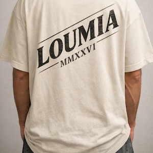 Op de afbeelding: Crèmekleurig T-shirt met het woord "LOUMIA" in zwart diagonaal op de rug gedrukt, met "MMXXVI" eronder. Het shirt heeft een relaxte pasvorm en korte mouwen.