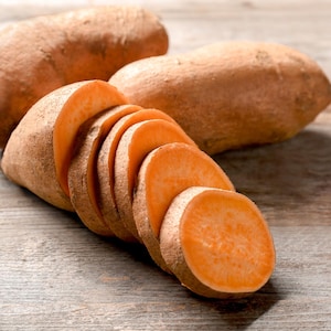 May include: Close-up of sweet potatoes, one sliced into rounds, with two whole sweet potatoes in the background. The sweet potatoes have a light brown skin and bright orange flesh. The image is set on a rustic wooden surface.