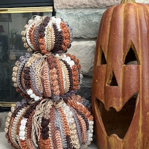 May include: A stack of three crocheted pumpkins in varying sizes, featuring vertical stripes of brown, tan, cream, and grey yarn with textured bobble stitches. A large, tall, carved orange jack-o'-lantern sits to the right of the pumpkins.