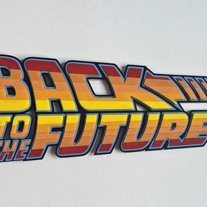 May include: A colourful sign featuring the text "BACK TO THE FUTURE" in a stylised font. The letters are in shades of yellow, orange, and red, outlined in blue and black. The sign is a retro design.