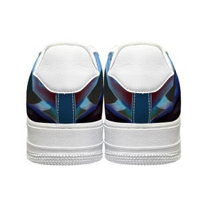 Op de afbeelding: Paar witte sneakers met een kleurrijk geometrisch ontwerp op de hiel. Het ontwerp heeft tinten blauw, rood en zwart. De schoenen hebben witte zolen en witte hielstukken.