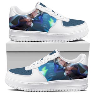 Op de afbeelding: Witte en blauwe sneakers met een afbeelding van een vrouw in een blauwe outfit. De sneakers hebben een witte zool en veters, met een blauwe neus en hiel. De afbeelding is van een vrouw met blond haar.