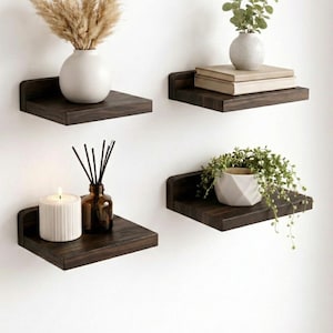 Peut inclure: Quatre étagères flottantes en bois brun foncé fixées à un mur blanc. Chaque étagère présente des objets décoratifs : un vase avec des plantes séchées, des livres, un petit vase avec de la verdure, une bougie et un flacon diffuseur.