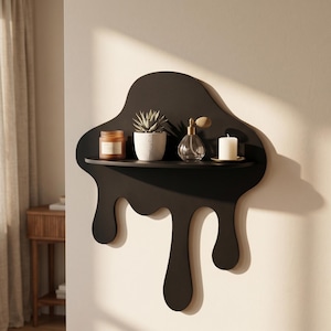 May include: A matte black wall-mounted shelf with a fluid, dripping paint design. The shelf holds a brown glass candle jar, a small white textured succulent pot, a clear glass perfume bottle with a gold atomizer, and a white pillar candle.
