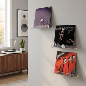 Op de afbeelding: Drie transparante acryl planken voor platen, gemonteerd aan een grijze muur, elk met een vinylplaat. De platen hebben albumhoezen met verschillende kleuren en ontwerpen, waaronder een rood-zwarte hoes en een zwart-wit portret.